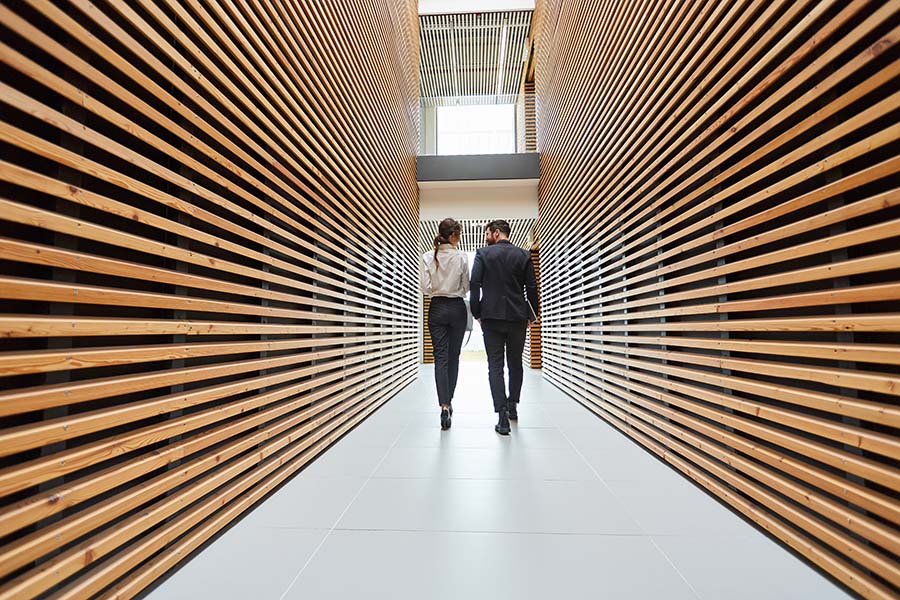 About Our Agency - Rear View of Two Business Colleagues Walking Down a Hallway in a Modern Office with Wooden Panels on the Walls